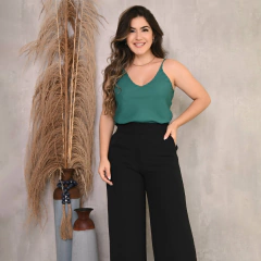 Mulher sorrindo, usando blusa verde e calça preta, posando ao lado de plantas decorativas.