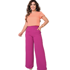 Mulher sorridente vestindo blusa laranja e calça roxa, posando de maneira confiante.