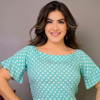 Mulher sorridente usando uma blusa verde com bolinhas brancas, com cabelos soltos e brincos. Fundo cinza.