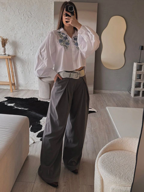 Modelo con camisa Catalina blanca, de corte corto, mangas abullonadas y bordado floral azul en el frente, combinada con pantalón gris y cinturón plateado
