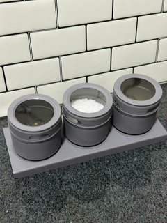Conjunto de tres especieros imantados grises con tapa transparente, organizados sobre una base en una encimera de cocina con fondo de azulejos blancos.