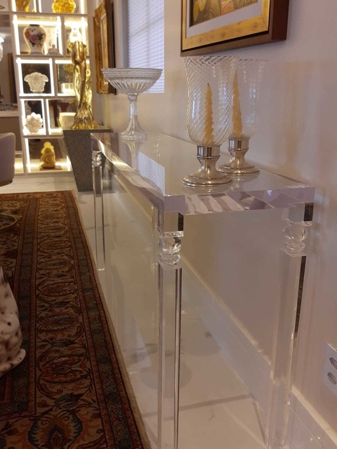 Aparador lapidário transparente de acrílico com detalhes clássicos, decorado com castiçais de vidro e vela, em ambiente interno elegante com tapete e obras de arte.