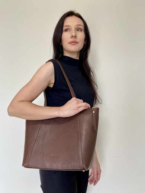 Mulher segurando uma bolsa tote marrom chamada Luna, com uma blusa preta e um fundo neutro.