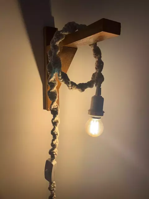 Luminária de macramê com suporte de madeira e lâmpada acesa, exibindo um design artesanal e elegante.