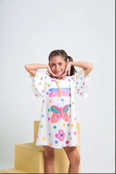 Criança sorridente vestindo um vestido branco com estampas coloridas de borboletas e pontos.