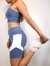 Modelo vestindo top azul e shorts de treino, alongando a perna em um ambiente claro.