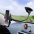 Suporte celular articulado com trava automática instalado no painel de um carro, segurando um smartphone, com fundo de estrada e campo.