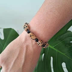 Pulseira de Jaspe Dálmata com pedras de Turmalina Negra, vista no pulso de uma pessoa, com fundo de folhas verdes.