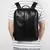 Mochila masculina de couro preta retrô multifuncional, sendo usada por um homem com camisa com listras horizontais cinza e branco, ideal para transporte seguro de notebook de 15.6 polegadas.