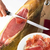 Um presunto cru jamón espanhol serrano sendo fatiado com uma faca, próximo a um prato com fatias do presunto.