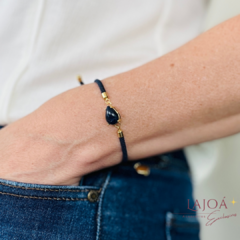 Pulseira cordão com pingente em forma de gota, em tom escuro, usada no pulso de uma pessoa.