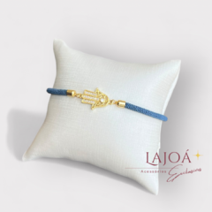 Pulseira Mão de Fátima em ouro com detalhe azul, apresentada sobre uma almofada branca.