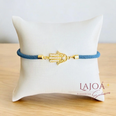 Pulseira Mão de Fátima em ouro com detalhe em azul, exibida sobre um suporte acolchoado.