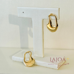 Brinco argola orgânica pequeno em banho de ouro, exibido em suporte branco.