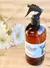 Aromatizador en spray para telas "Petite Affaire" con tapa negra y etiqueta blanca, junto a flores blancas.
