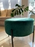 Sillón puff Marlon de color verde, con patas negras, ubicado en un ambiente luminoso. Se puede ver una planta y un sofá en el fondo.