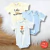 Kit de 3 bodies para bebê, com duas peças estampadas e uma lisa, em cores azul claro, bege com frase "little cute elephant" e branco com estampa de elefante segurando balão laranja