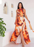Modelo usando um vestido longo com estampa em tons de fogo, destacando recortes e fenda, em ambiente claro.
