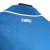 Camisa titular Napoli 2024-2025 azul celeste com mosaico de escudos em embossing. Gola pólo azul com contorno branco e punhos brancos. Nome do time na nuca. Escudo branco no peito esquerdo, logo EA7 branco no direito.
