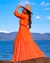 Modelo usando uma saída de praia longa laranja com um chapéu de palha, posando à beira do mar.
