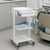 White multifunctional dental trolley with drawer and two shelves on wheels next to a dental chair in a clinic setting