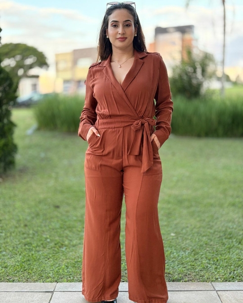 Mulher usando um macacão terra com manga longa e cinto, em um ambiente externo com grama ao fundo.