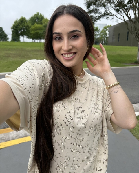 Jovem mulher sorrindo e posando com uma blusa Hotfix GLAM em um ambiente externo, com árvores ao fundo.