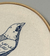 Detalhe do bastidor bordado com a ilustração de um pássaro saíra-sapucaia em um fundo claro.