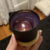 Vela aromática de cera de coco na cor preta com chama acesa, em um suporte de madeira.