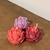 Três velas aromáticas em forma de flor de lótus, nas cores roxa, vermelha e rosa, sobre uma superfície de madeira.