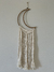 Filtro decorativo de macramê com padrão de tranças nas franjas, em formato de lua crescente, pendurado em parede branca, feito artesanalmente em fios de algodão cru.