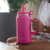 Vaso SoloVasos Pro de color rosa con tapa y sorbete, sostenido por una persona con pulseras, sobre sus piernas en un ambiente interior con plantas.