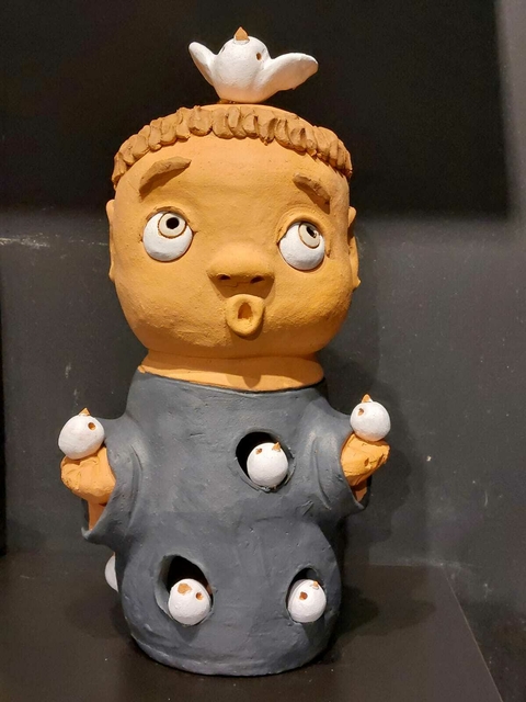 Escultura de cerâmica de uma figura com expressão surpresa, com um passarinho branco em cima da cabeça e outros passarinhos em aberturas ao redor do corpo.