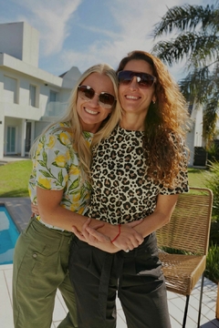 Dos mujeres sonrientes abrazadas, una con remera de estampado de limones y la otra con remera de estampado de leopardo, en un entorno al aire libre con piscina y palmeras.