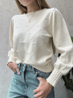 Una mujer usa un suéter blanco llamado Nube, con detalles en relieve, combinado con jeans de tiro alto.
