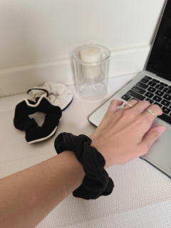 Una mano con un scrunchie negro en la muñeca, junto a otros scrunchies y una vela sobre una mesa con una computadora portátil.