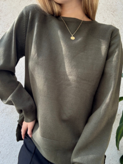 Un suéter de color verde oliva, con detalles en el tejido, usado por una persona que sostiene una mano en la cadera. Se destaca un collar dorado en el cuello.