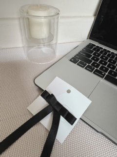 Prensita Moño negra sobre una superficie clara, junto a una vela blanca y una laptop.