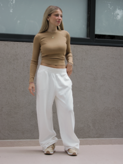 Mujer de pie luciendo un pantalón blanco llamado Neblina, con un top de manga larga beige y zapatillas claras, en un entorno moderno.