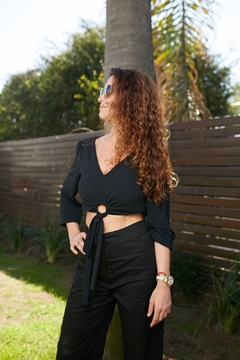 Modelo luciendo una blusa negra con un nudo en el centro y pantalones oscuros, en un ambiente exterior con palmeras de fondo.