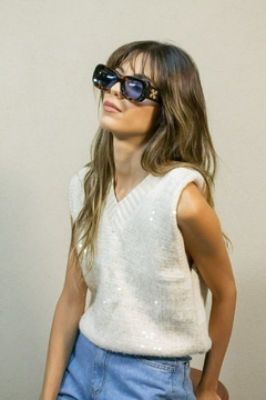 Mujer usando chaleco blanco sin mangas con detalles de lentejuelas y jeans celestes, llevando gafas de sol grandes y oscuras