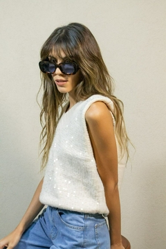 Mujer usando chaleco blanco sin mangas con detalles de lentejuelas y jeans celestes, llevando gafas de sol grandes y oscuras