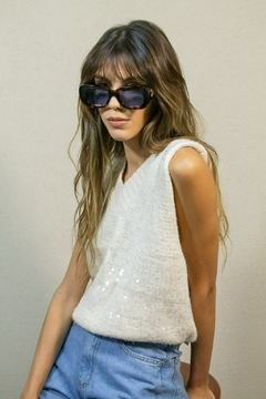 Mujer usando chaleco blanco sin mangas con detalles de lentejuelas y jeans celestes, llevando gafas de sol grandes y oscuras