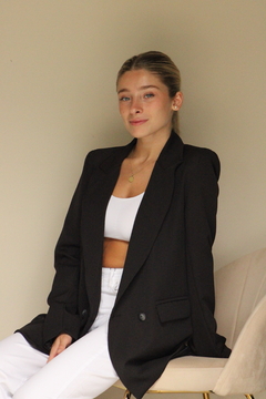 Mujer sentada en una silla con un saco negro sobre una blusa blanca y pantalones blancos, sonriendo frente a una pared clara.