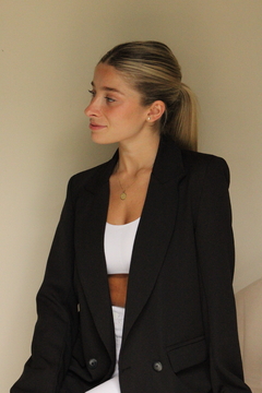 Mujer con un saco malaquita negro, sentada en un ambiente neutro, usando un top blanco y con cabello lacio y recogido.