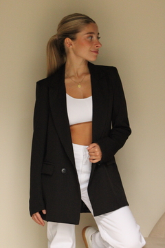 Mujer luciendo un saco malaquita negro, combinando con un top blanco y pantalones blancos, en un fondo neutro.