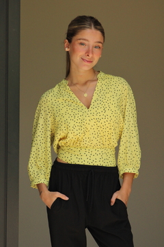 Modelo luciendo una blusa amarilla con estampado negro, de manga larga y escote en V, combinada con pantalones negros.