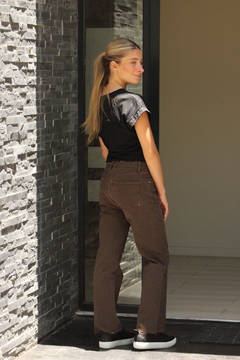 Mujer de espaldas usando una remera negra y pantalones marrones, posando frente a una pared de piedra.