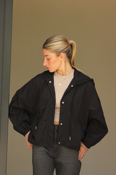 Mujer luciendo un rompeviento negro con capucha, combinado con una camiseta clara y jeans oscuros, en un entorno minimalista.