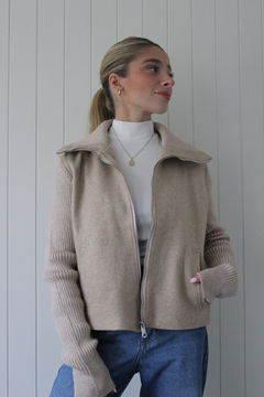 Campera Marea de color beige, con cierre frontal y mangas de punto, combinada con una camiseta blanca y jeans, exhibida en un entorno minimalista.